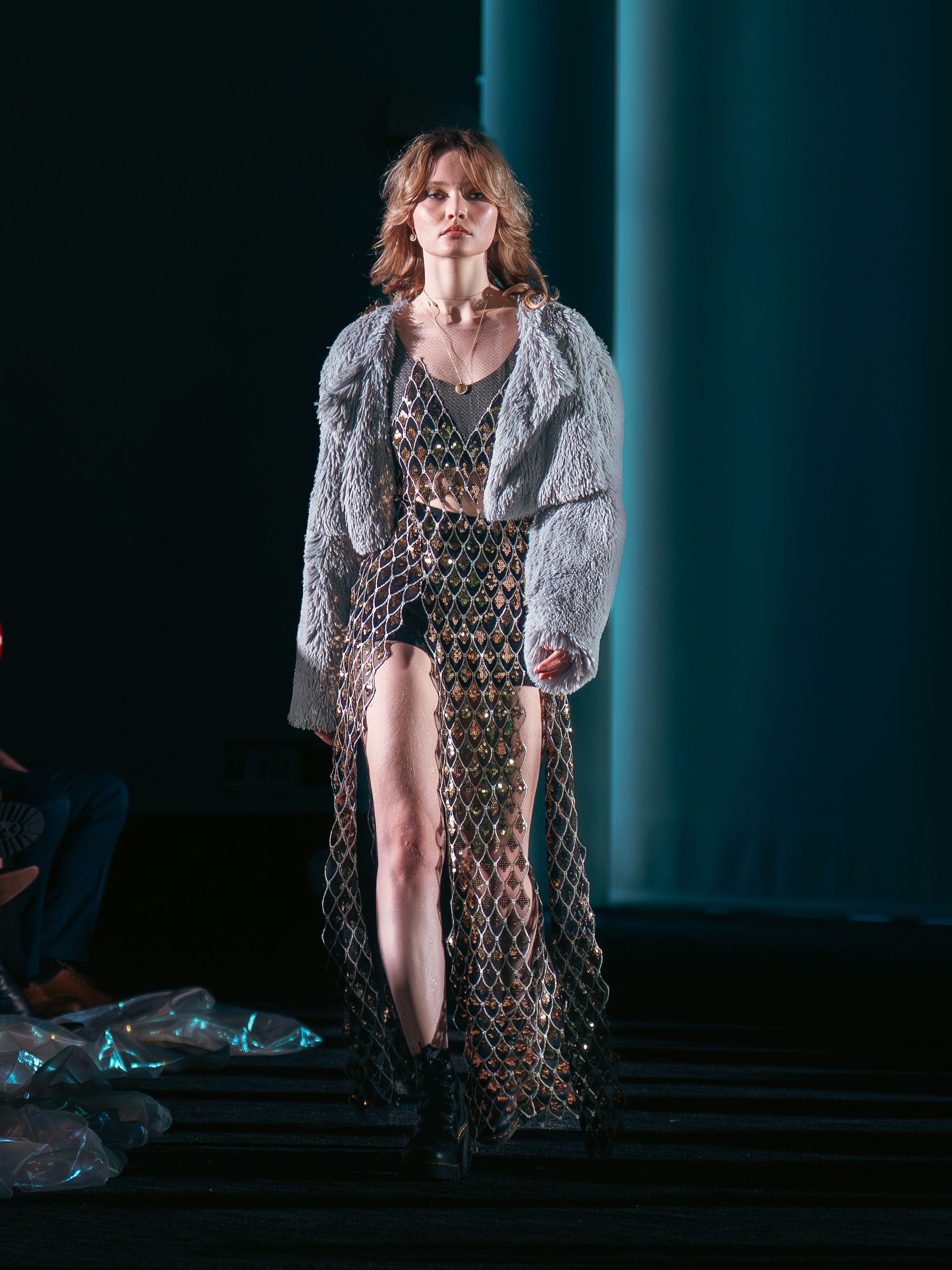 Model walks down the runway in a textured gray faux fur jacket over a sheer metallic dress made of interlinked, reflective diamond-shaped pieces. She wears black boots and gold jewelry.